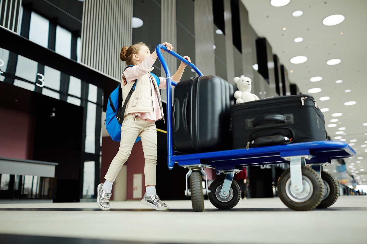 Little traveler with backpack pushing baggage cart with large suitcases and toy in airport lounge